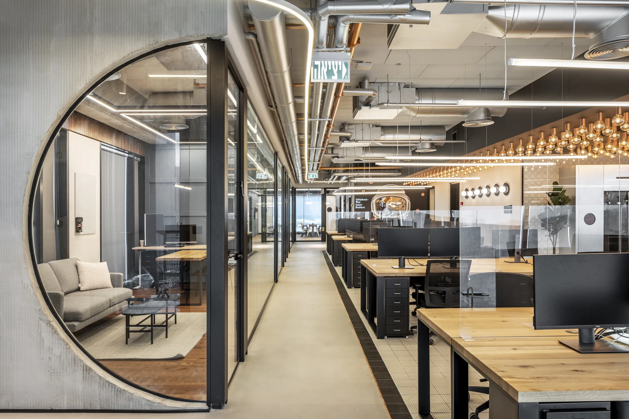 An open-plan workspace with wood desks and glass pods facing the Edison-bulb canopy